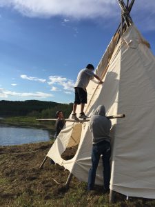 Two people set up a teepee on the shores of a lake.