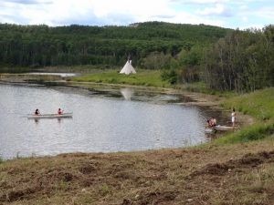 Two people paddle a canoe in the middle of a lake, as seen from the shore. A teepee can be seen in the background.