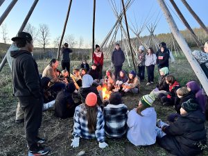 A group of about 20 youth sit around a fire, underneath tree trunks shaped like a teepee, listening to an elder speak.