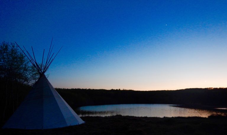 The sun sets on a small lake surrounded by a forest, as seen from beside a teepee.