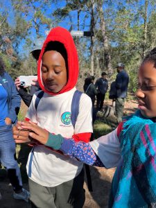 Out on a forest trail, a young boy holds a small snake in his hands, while his sister, standing to his left, reaches out a hand to touch the snake.